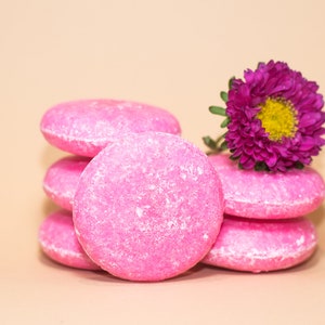 May include: A stack of five pink solid shampoo bars with a purple flower on top. The shampoo bars are round and have a textured surface.