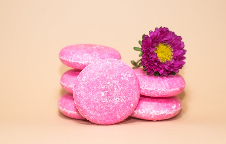 May include: A stack of five pink solid shampoo bars with a purple flower on top. The shampoo bars are round and have a textured surface.