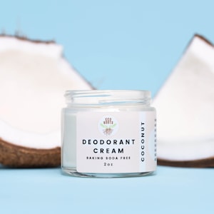 May include: A white jar with a green logo that says "Eco Roots" and the text "Deodorant Cream Coconut Baking Soda Free 2 oz". The jar is sitting on a light blue surface with two halves of a coconut on either side.