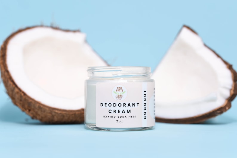 May include: A white jar with a green logo that says "Eco Roots" and the text "Deodorant Cream Coconut Baking Soda Free 2 oz". The jar is sitting on a light blue surface with two halves of a coconut on either side.