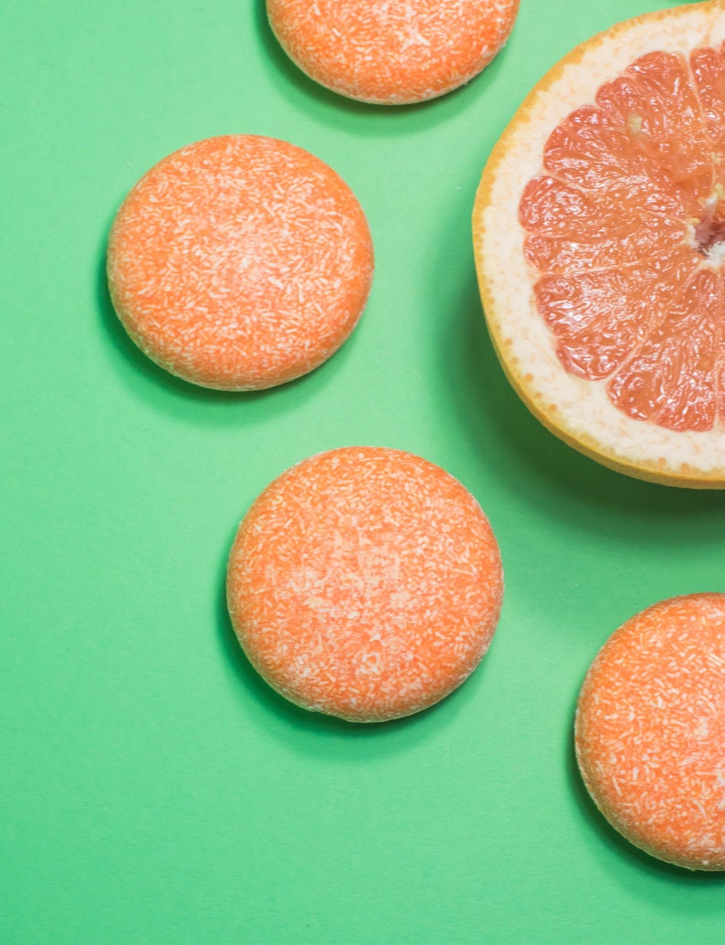 May include: Four round orange and white solid shampoo bars and a half grapefruit on a green background.