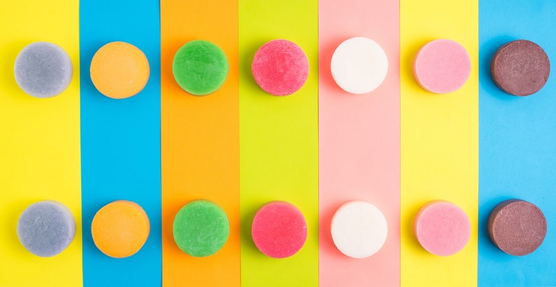 Pode incluir: Uma fila de dez barras de sab&atilde;o redondas e coloridas dispostas sobre um fundo listrado. As cores das barras de sab&atilde;o s&atilde;o cinza, laranja, verde, rosa, branco, rosa, marrom, cinza, laranja, verde, rosa, branco, rosa, marrom.