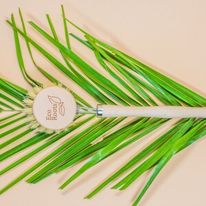 May include: A wooden dish brush with a natural bristle head and a wooden handle. The brush has a round wooden tag with the text "Eco Roots" and a leaf design.
