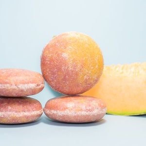 May include: Four round pink and orange soap bars stacked on top of each other, with a slice of cantaloupe melon to the right.
