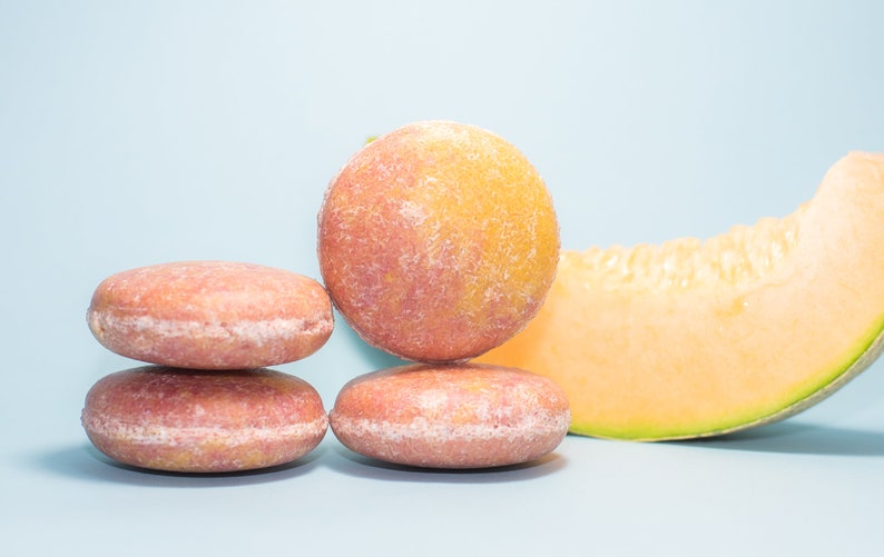 May include: Four round pink and orange soap bars stacked on top of each other, with a slice of cantaloupe melon to the right.