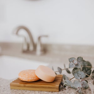 May include: Two round bars of shampoo soap, one orange and one light pink, are on a wooden soap dish. A sprig of green eucalyptus is in the background.