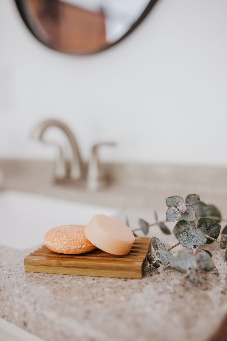 May include: Two round bars of shampoo soap, one orange and one light pink, are on a wooden soap dish. A sprig of green eucalyptus is in the background.