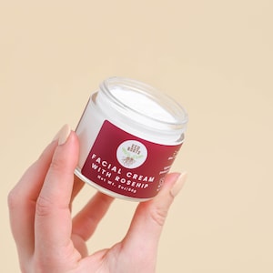 May include: A white jar with a red and white label that reads "Facial Cream with Rosehip" and "Eco Roots". The jar is being held by a hand.