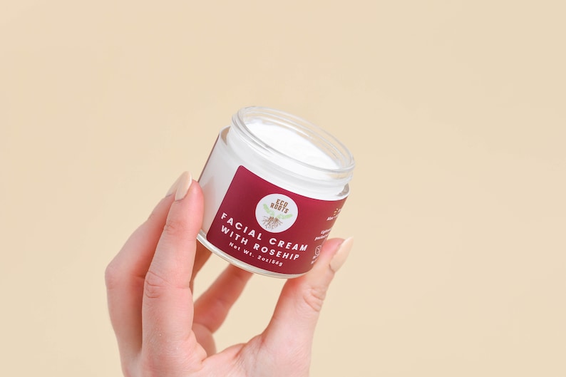 May include: A white jar with a red and white label that reads "Facial Cream with Rosehip" and "Eco Roots". The jar is being held by a hand.