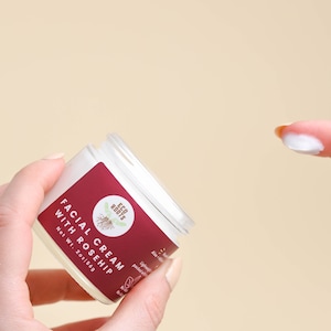 May include: A white jar of facial cream with a red label that says "Facial Cream with Rosehip" and "Eco Roots". The jar is being held in a hand, and a finger is shown with a dollop of cream on it.