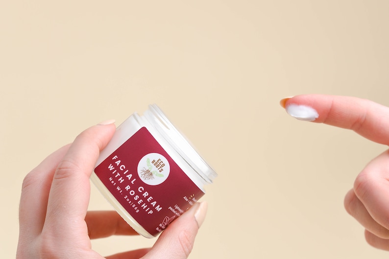 May include: A white jar of facial cream with a red label that says "Facial Cream with Rosehip" and "Eco Roots". The jar is being held in a hand, and a finger is shown with a dollop of cream on it.