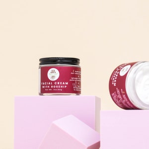 May include: Two glass jars of facial cream with black lids. The jar on the left is labeled "Facial Cream with Rosehip" and is sitting on a pink platform. The jar on the right is lying on its side on a pink platform.