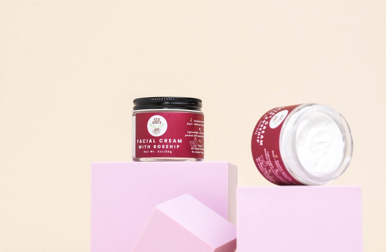 May include: Two glass jars of facial cream with black lids. The jar on the left is labeled "Facial Cream with Rosehip" and is sitting on a pink platform. The jar on the right is lying on its side on a pink platform.