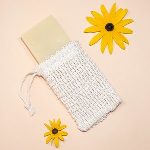 May include: A white mesh bag with a drawstring closure, containing a rectangular bar of soap. Two yellow flowers with dark centers are on a light pink background.