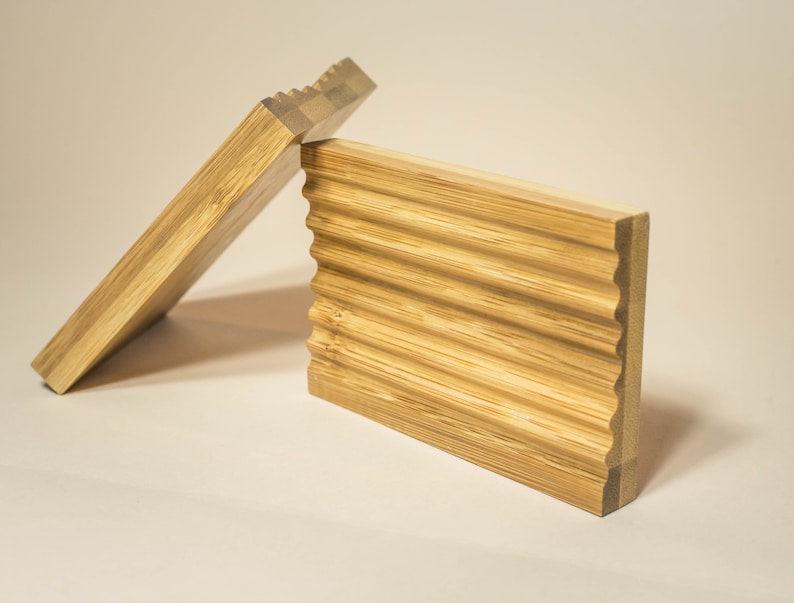 May include: Two wooden soap dishes made of bamboo. The top dish has a wavy design and the bottom dish is flat.