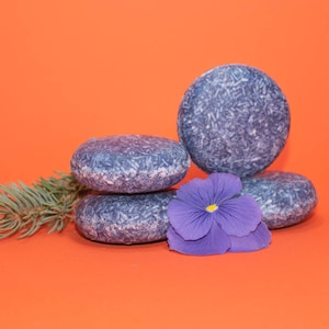 May include: Four blue shampoo bars stacked on top of each other on an orange background. A purple flower is in front of the bars.