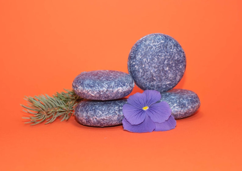 May include: Four blue shampoo bars stacked on top of each other on an orange background. A purple flower is in front of the bars.