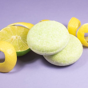 May include: Two green and white solid shampoo bars with a lime wedge and lemon peel on a purple background.