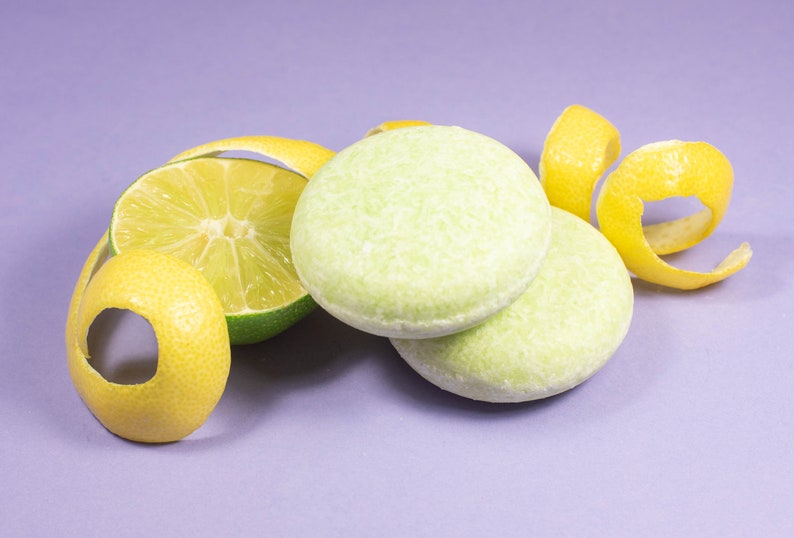 May include: Two green and white solid shampoo bars with a lime wedge and lemon peel on a purple background.