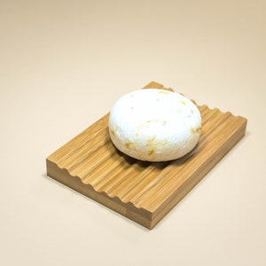 May include: A white round bar of soap with brown flecks sits on a wooden soap dish with a wavy edge.