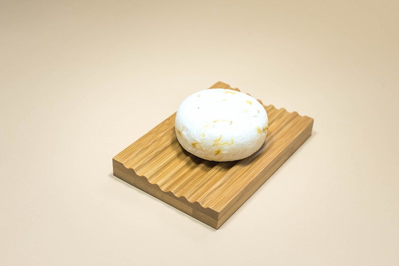 May include: A white round bar of soap with brown flecks sits on a wooden soap dish with a wavy edge.