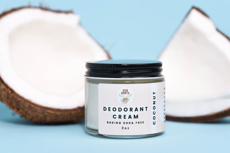 May include: A glass jar of coconut deodorant cream with a black lid. The jar is labeled "ECO ROOTS DEODORANT CREAM BAKING SODA FREE 2 oz COCONUT". The jar is sitting on a light blue surface with two halves of a coconut behind it.