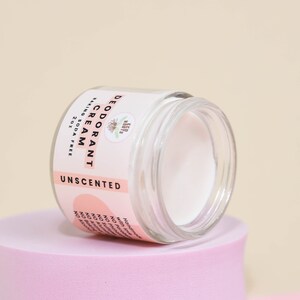May include: A white, unscented deodorant cream in a glass jar with a pink label that reads "DEODORANT CREAM, BAKING SODA FREE, 2 OZ, UNSCENTED".