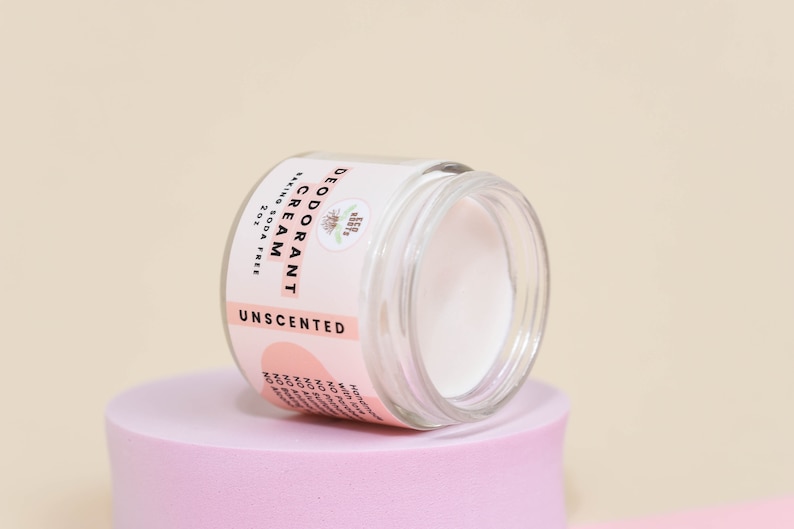 May include: A white, unscented deodorant cream in a glass jar with a pink label that reads "DEODORANT CREAM, BAKING SODA FREE, 2 OZ, UNSCENTED".