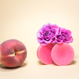 Pode incluir: Dois sabonetes redondos cor-de-rosa com um grupo de rosas roxas no topo. Os sabonetes est&atilde;o sobre uma superf&iacute;cie bege claro.