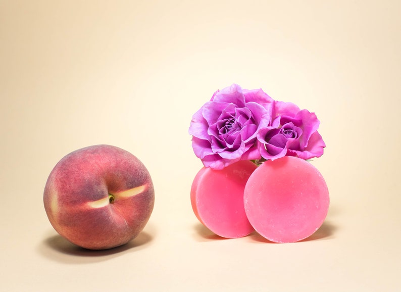 Pode incluir: Dois sabonetes redondos cor-de-rosa com um grupo de rosas roxas no topo. Os sabonetes est&atilde;o sobre uma superf&iacute;cie bege claro.