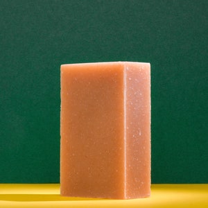May include: A rectangular bar of soap with a light brown color and a rough texture. The soap is sitting on a yellow surface in front of a green background.
