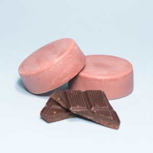 Pode incluir: Duas barras de sab&atilde;o redondas cor-de-rosa e tr&ecirc;s peda&ccedil;os de chocolate preto sobre um fundo azul claro.