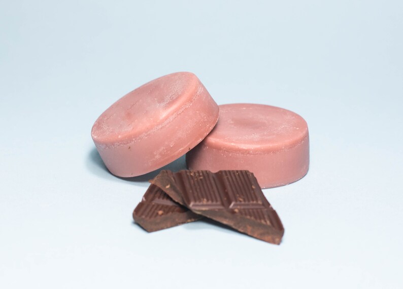 Pode incluir: Duas barras de sab&atilde;o redondas cor-de-rosa e tr&ecirc;s peda&ccedil;os de chocolate preto sobre um fundo azul claro.