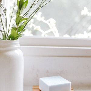 May include: A white bar of soap sits on a bamboo soap dish. A white vase with green stems and purple flowers sits on a white countertop.