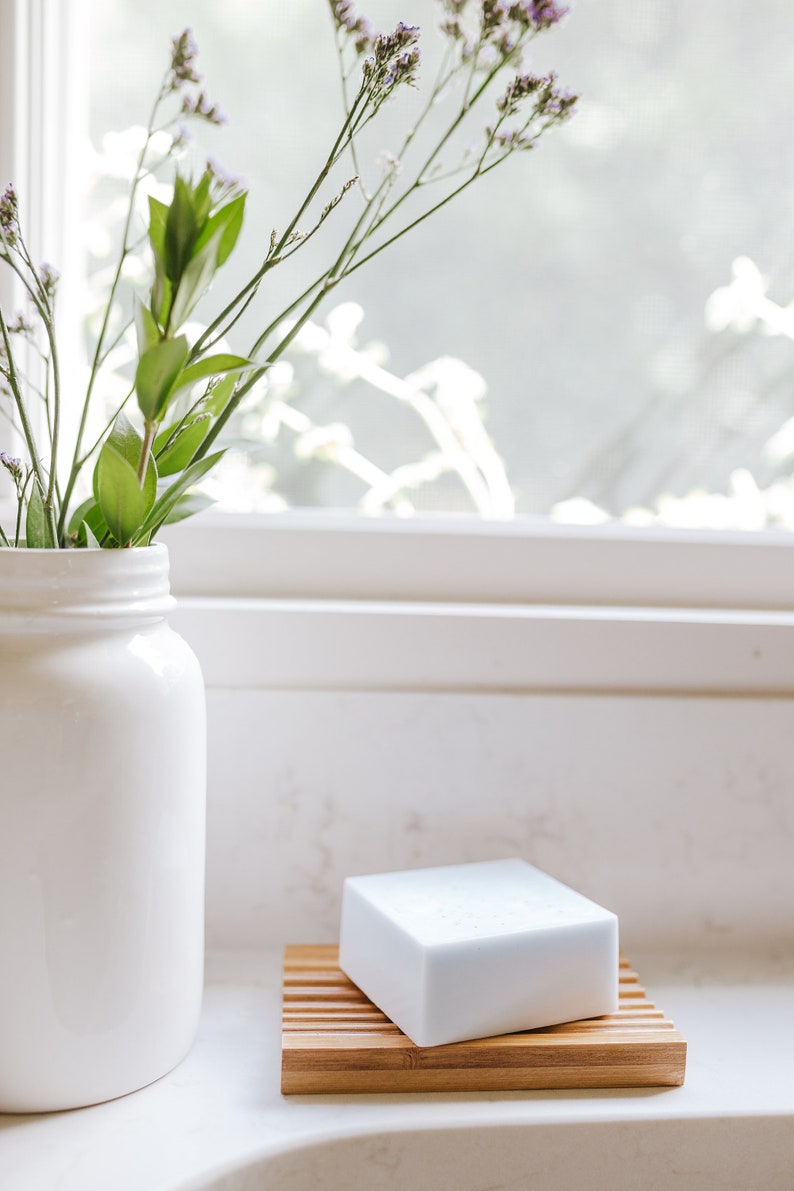 May include: A white bar of soap sits on a bamboo soap dish. A white vase with green stems and purple flowers sits on a white countertop.