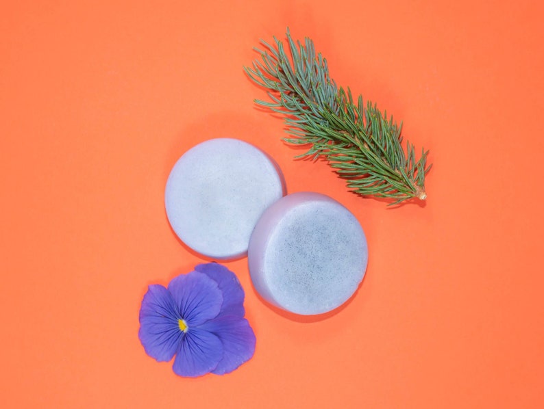 Pode incluir: Duas barras de sab&atilde;o redondas de cor cinza claro sobre um fundo laranja brilhante. Tamb&eacute;m h&aacute; um ramo de agulhas de pinho verdes e uma flor de pensamento p&uacute;rpura no fundo.