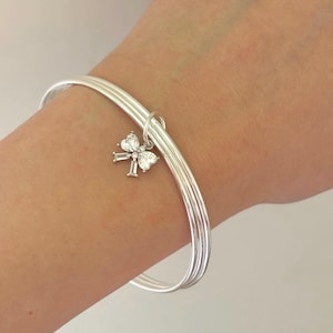 May include: A silver bangle bracelet with three bands. A small charm in the shape of a bow with clear, sparkling stones hangs from the bracelet. The bracelet is on a person's wrist.