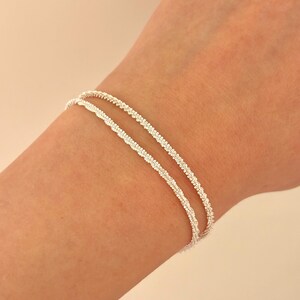 May include: A delicate double-strand bracelet featuring a fine, textured silver chain. The two thin, shimmering strands wrap around the wrist, creating a layered appearance with a consistent, intricate link pattern throughout the entire design.