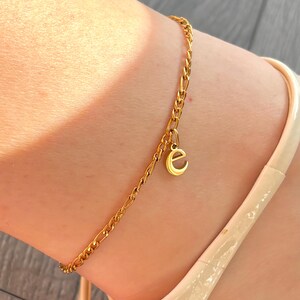 May include: A gold-colored anklet with a chain link design and a small charm in the shape of the letter "e". The anklet is worn on the ankle. The anklet is made of a shiny, reflective material.