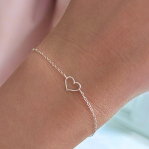 May include: A fine silver bracelet with a heart-shaped pendant. The heart is a simple outline, suspended from a delicate chain. The bracelet is shown on a wrist, highlighting its minimalist design and elegant style. The chain is made of small, linked segments.