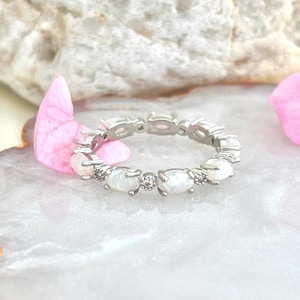 Könnte beinhalten: Ein silberner Ring mit ovalen, schillernden Edelsteinen und kleinen Akzentsteinen. Der Ring liegt auf einer weiß-grauen Oberfläche, mit rosa Blütenblättern im Hintergrund. Der Ring ist ein Schmuckstück.