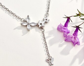May include: A silver-toned necklace featuring a delicate chain with a central pendant design. The pendant consists of a four-petaled flower shape with clear, marquise-cut stones, two small metal flowers, and a dangling chain with a butterfly charm.