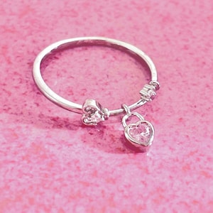 Peut inclure: Une bague en argent avec un charm en forme de cadenas en forme de cœur. La bague a un design délicat avec un petit nœud et quelques petits cristaux. La bague est posée sur un fond rose.
