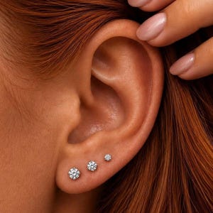 May include: Three round, clear, faceted stud earrings in graduated sizes, arranged in a row along the earlobe. Each earring features a brilliant-cut stone set in a metallic prong setting, creating a sparkling, reflective appearance against the skin.
