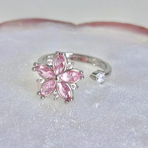 Può includere: Un anello regolabile color argento con un design a fiore rosa. Il fiore ha sei petali rosa e piccoli accenti bianchi. Un'unica gemma trasparente è incastonata sull'estremità aperta dell'anello. L'anello è posto su uno sfondo bianco.