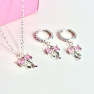 May include: A silver necklace and earring set featuring pink bow-shaped pendants. The necklace has a delicate chain, and the earrings are hoop style. The bows are adorned with pink gemstones. The jewelry set is displayed on a white surface with a pink background.