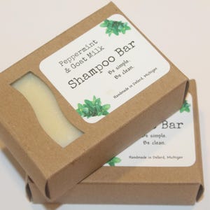 Op de afbeelding: Twee bruine kartonnen dozen met witte etiketten. De bovenste doos heeft een etiket met de tekst "Peppermint & Goat Milk Shampoo Bar Be simple. Be clean. Handmade in Deford, Michigan". De onderste doos heeft een etiket met de tekst "Bar Be simple. Be clean Handmade in Deford, Michigan".
