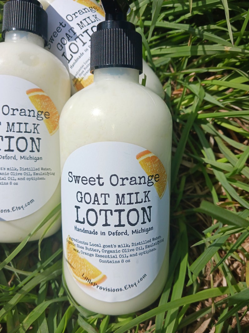 May include: A white bottle of Sweet Orange Goat Milk Lotion, handmade in Deford, Michigan. The bottle has a black pump top and a white label with orange text. The label lists the ingredients: local goat's milk, distilled water, shea butter, organic olive oil, emulsifying wax, orange essential oil, and optiphen. The bottle contains 8 ounces of lotion.
