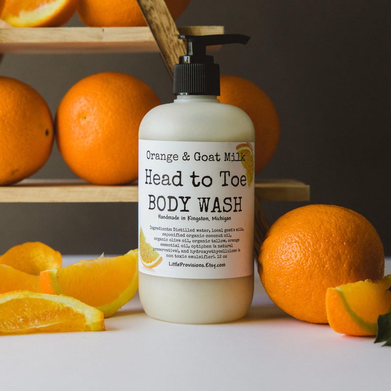 Op de afbeelding: Een witte fles met oranje- en geitenmelk-douchegel met een pompdispenser. De fles heeft een zwart etiket met de tekst "Orange & Goat Milk Head to Toe BODY WASH Handmade in Kingston, Michigan" en een lijst met ingredi&euml;nten. De fles staat op een houten plank met sinaasappelen eromheen.