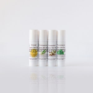 Könnte beinhalten: Vier weiße Tuben mit Bio-Hautbalsam mit Etiketten in verschiedenen Farben und Düften: Orange, Zitronengras, unparfümiert und Pfefferminze. Die Etiketten zeigen alle "Organic Skin Stick" und "ESTORE" in brauner Schrift.